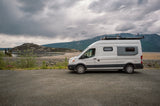White camper van (rv rental) parked on gravel, facing a scenic lake, mountains & cloudy sky. Ready for adventure!