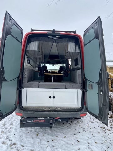 RV rental: Red camper van rear, doors open, showing converted interior with bed/storage, table, and hitch. Snowy ground.