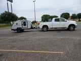 White pickup truck tows a compact Bushwhacker travel trailer, parked at a Walmart, an ideal RV rental.