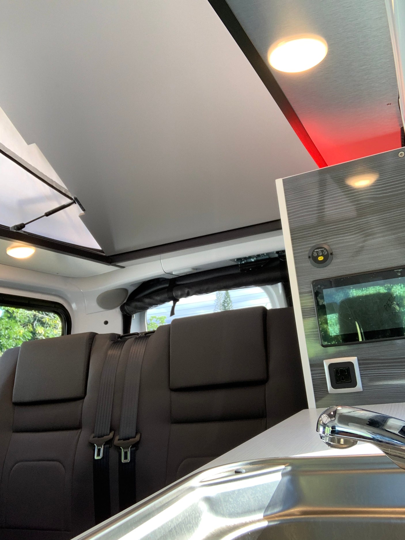 RV rental interior: Overhead cabinet with screen, power outlet, lighting. Comfy seating with seatbelts & a sink are also visible.