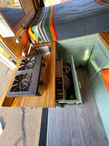 This RV rental features a compact kitchen with a stove, sink, and organized storage, plus a cozy bed with a colorful blanket.