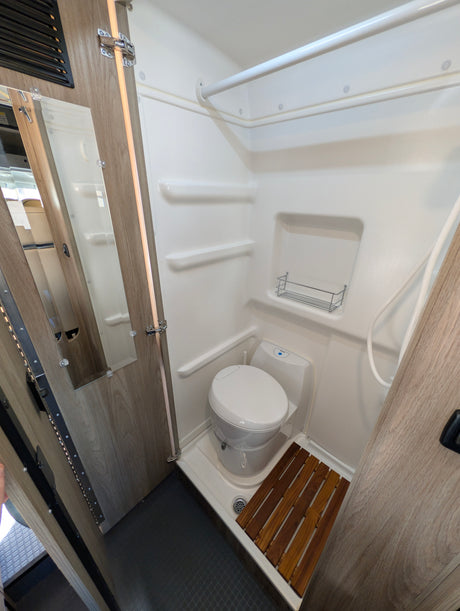 Inside an rv rental: a compact wet bath with a toilet, shower, built-in shelves, and a wooden slatted mat.