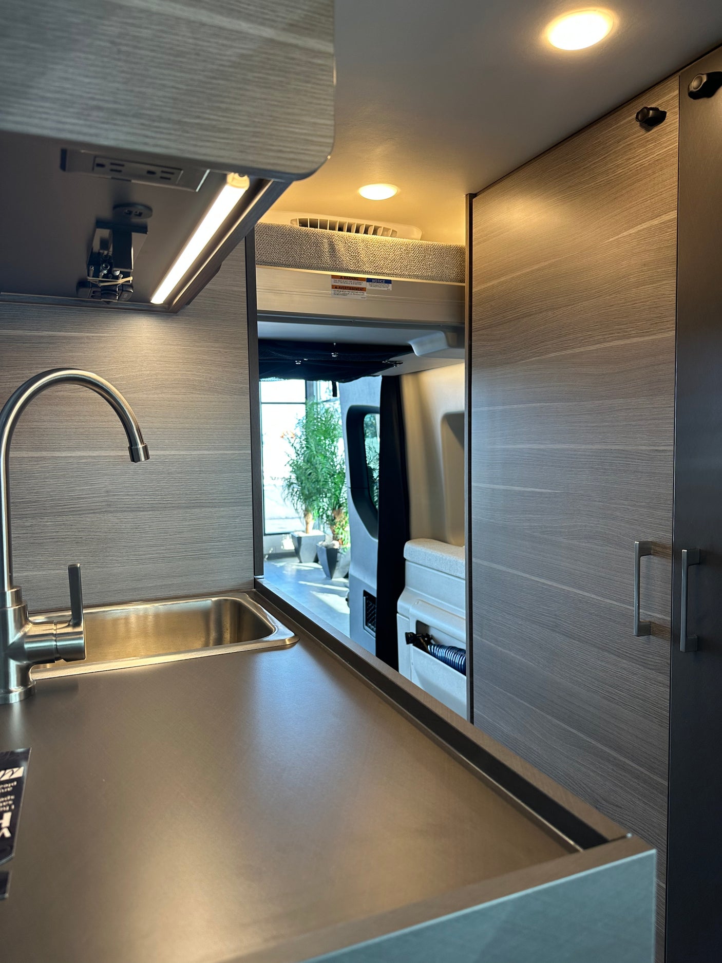 Modern rv rental interior: compact kitchen with a stainless steel sink, dark counter, LED lights, and a rear sleeping area.
