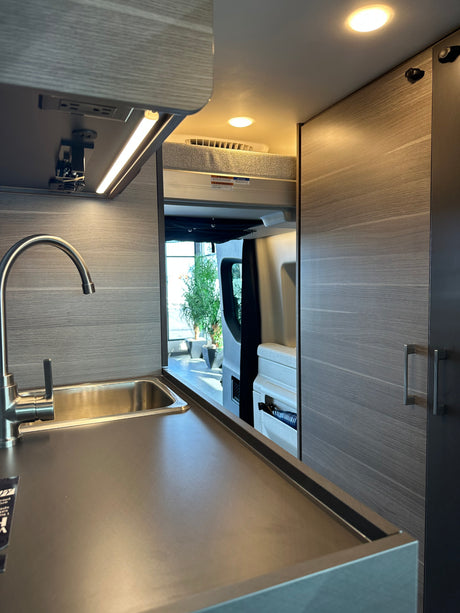 Modern rv rental interior: compact kitchen with a stainless steel sink, dark counter, LED lights, and a rear sleeping area.