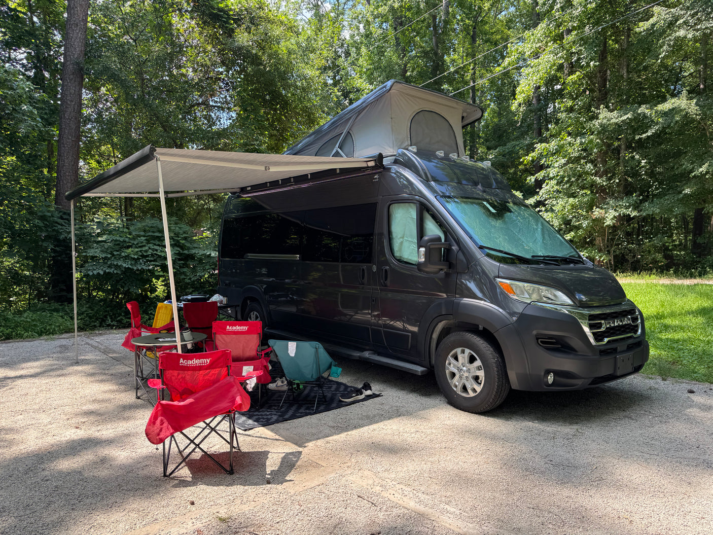 This RV rental, a dark gray camper van with a pop-top tent, is set up at a shady campsite with an awning, chairs, and table.