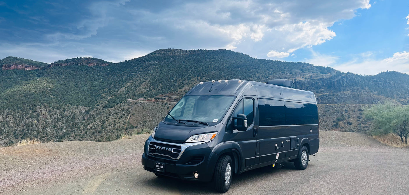 Dark gray Ram ProMaster camper van, an RV rental, parked on a dirt pull-off with rugged mountains & cloudy sky.
