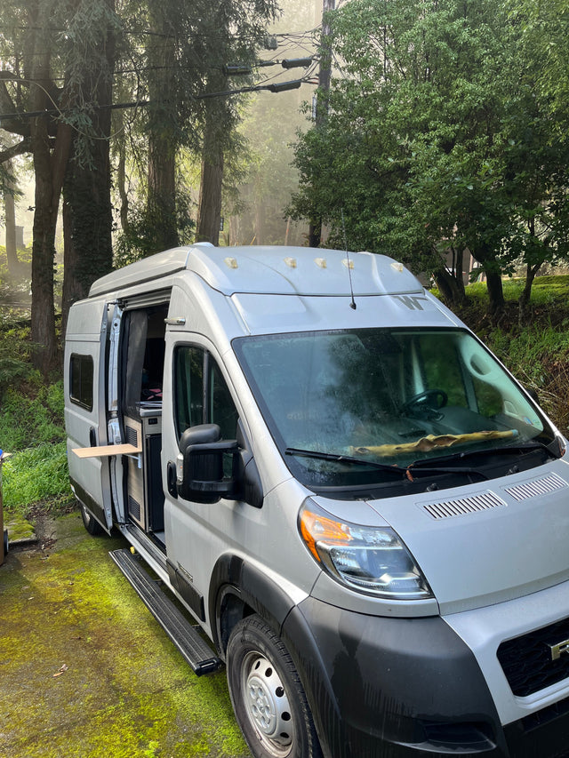 Discover adventure: a silver RV rental camper van parked amidst misty trees, with its side door open revealing the interior.
