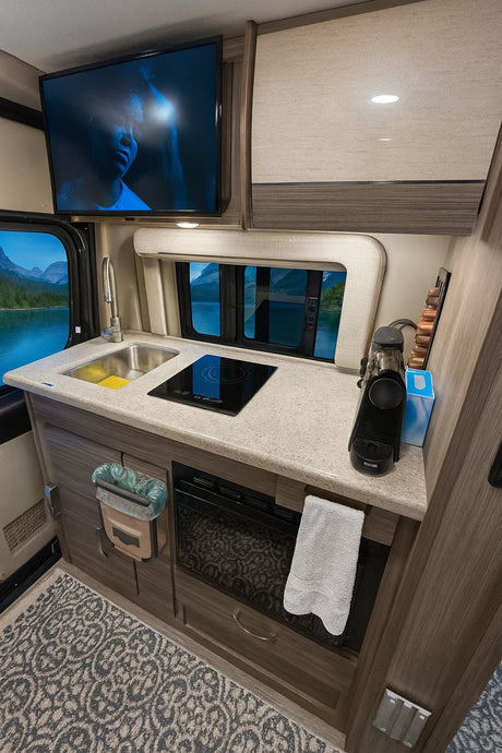 Compact rv rental kitchen featuring a TV, sink, induction cooktop, coffee maker, and windows displaying serene lake views.