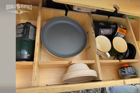 Organized RV rental storage with camping kitchen essentials: propane, pan, mugs, and plates in a wooden cubby.