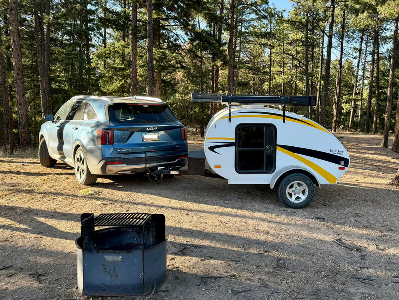 Blue SUV towing a white teardrop RV rental, parked at a wooded campsite. A fire pit is in the foreground, ready for camping. — photo 4