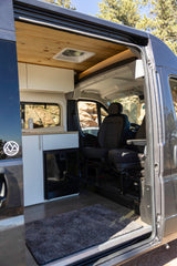 View inside an RV rental camper van: wooden ceiling, modern kitchen with fridge, front seats, & open side door revealing rocky views.
