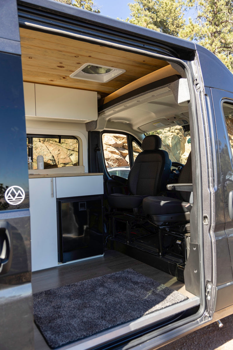 View inside an RV rental camper van: wooden ceiling, modern kitchen with fridge, front seats, & open side door revealing rocky views. — photo 11
