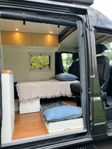 Spacious RV rental interior with bed, pull-out storage, wood cabinets, and front seats. View from open door.