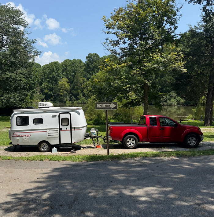 An rv rental, a white Scamp trailer towed by a red pickup truck, parked beside a lake surrounded by trees.