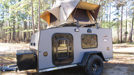 A rugged Escapade Camper Co. trailer with a pop-up rooftop tent sits in a forest; ideal for an RV rental getaway.