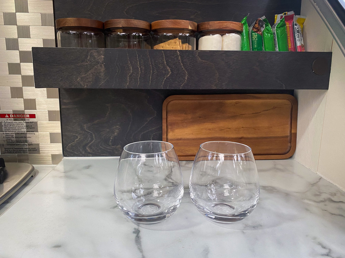 Inside an RV rental: Two glasses on a countertop, a shelf with snack jars, and a propane safety warning sign.