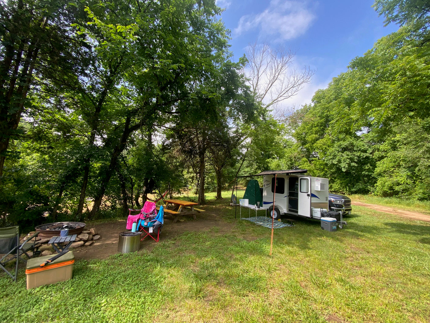 This peaceful rv rental campsite features a white camper, truck, picnic table, and fire pit, surrounded by lush green trees.