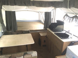 Bright pop-up camper interior, ideal for RV rental. Features bed, dining, & compact kitchen with stove & sink.