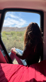 From an RV rental, a person with long hair views a sunny mountain landscape from inside, with red bedding in the foreground.
