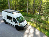 White camper van RV rental with roof rack and solar panels, parked on a sunlit forest road.
