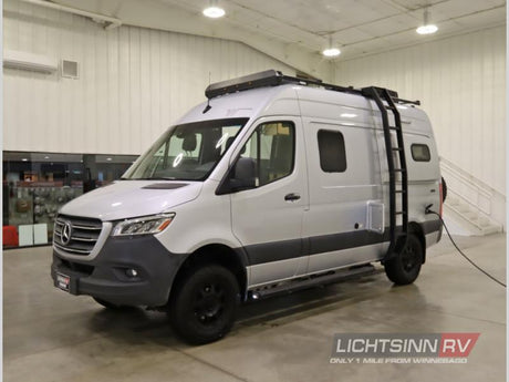 Silver Mercedes-Benz Sprinter camper van for rv rental, with rugged off-road tires and roof ladder, parked indoors.