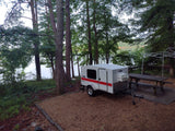 Small white & red teardrop RV rental at a serene lakeside campsite with trees & picnic table, perfect for outdoor adventures.