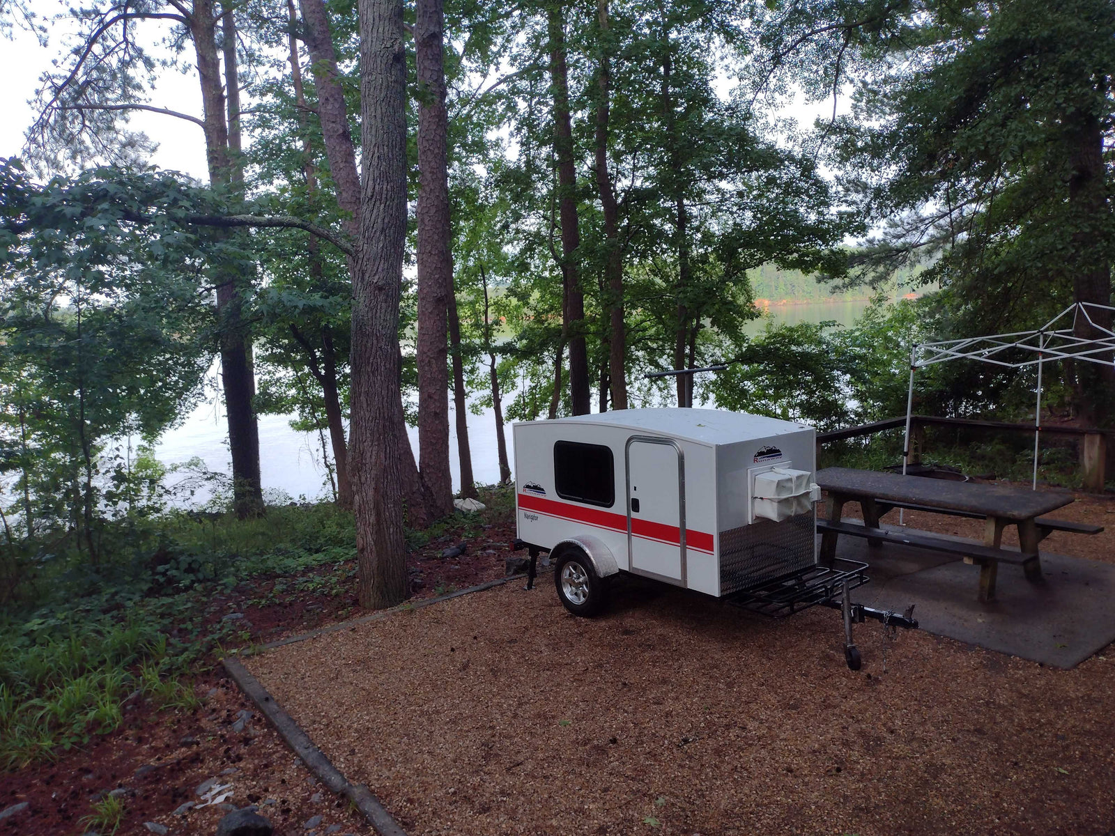 Small white & red teardrop RV rental at a serene lakeside campsite with trees & picnic table, perfect for outdoor adventures.