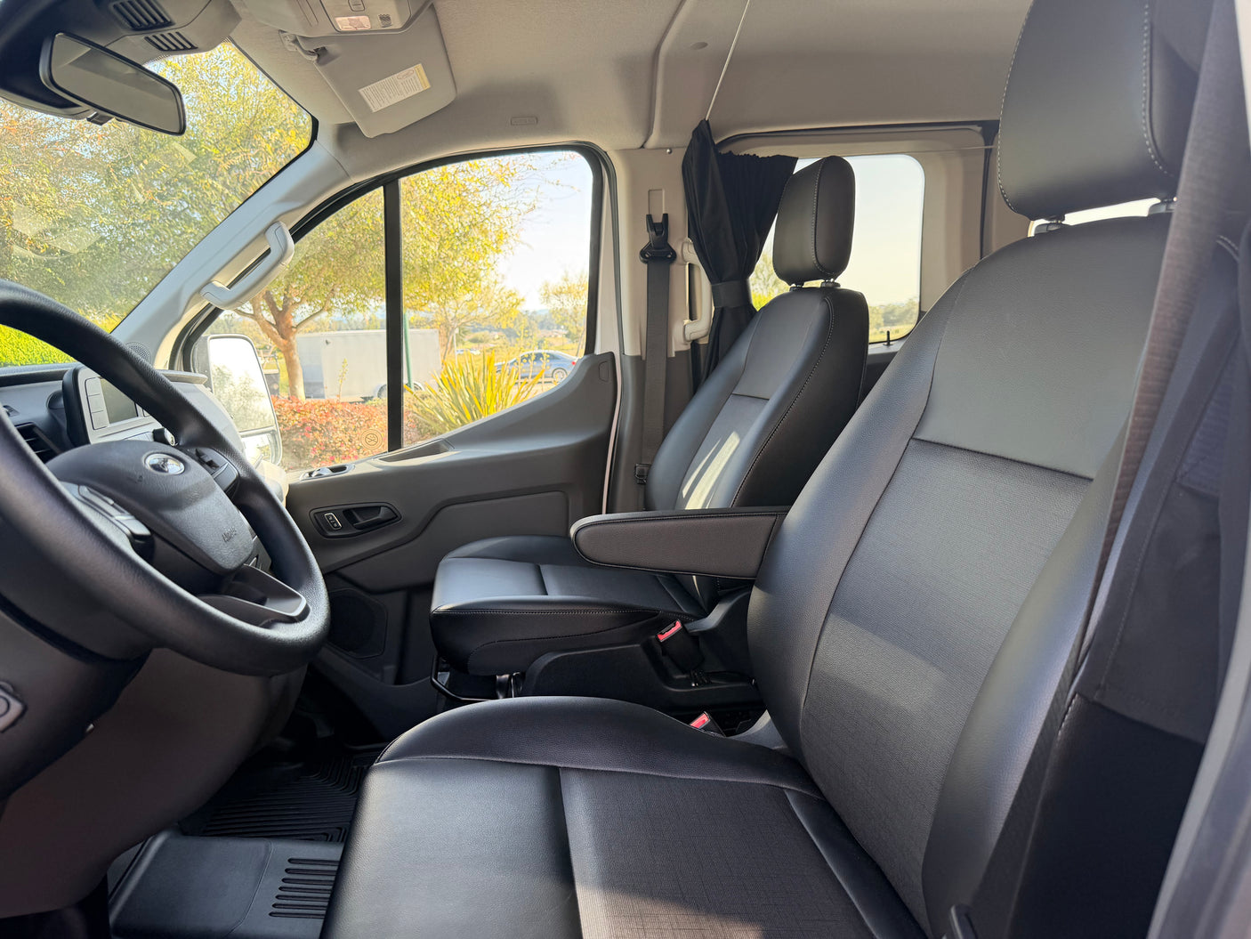 Front cabin of an RV rental, showing black leather seats, steering wheel, and a sunny view through the windshield.
