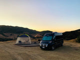 Black RV rental van and screen tent with people, set up at a scenic mountain camp during sunset.