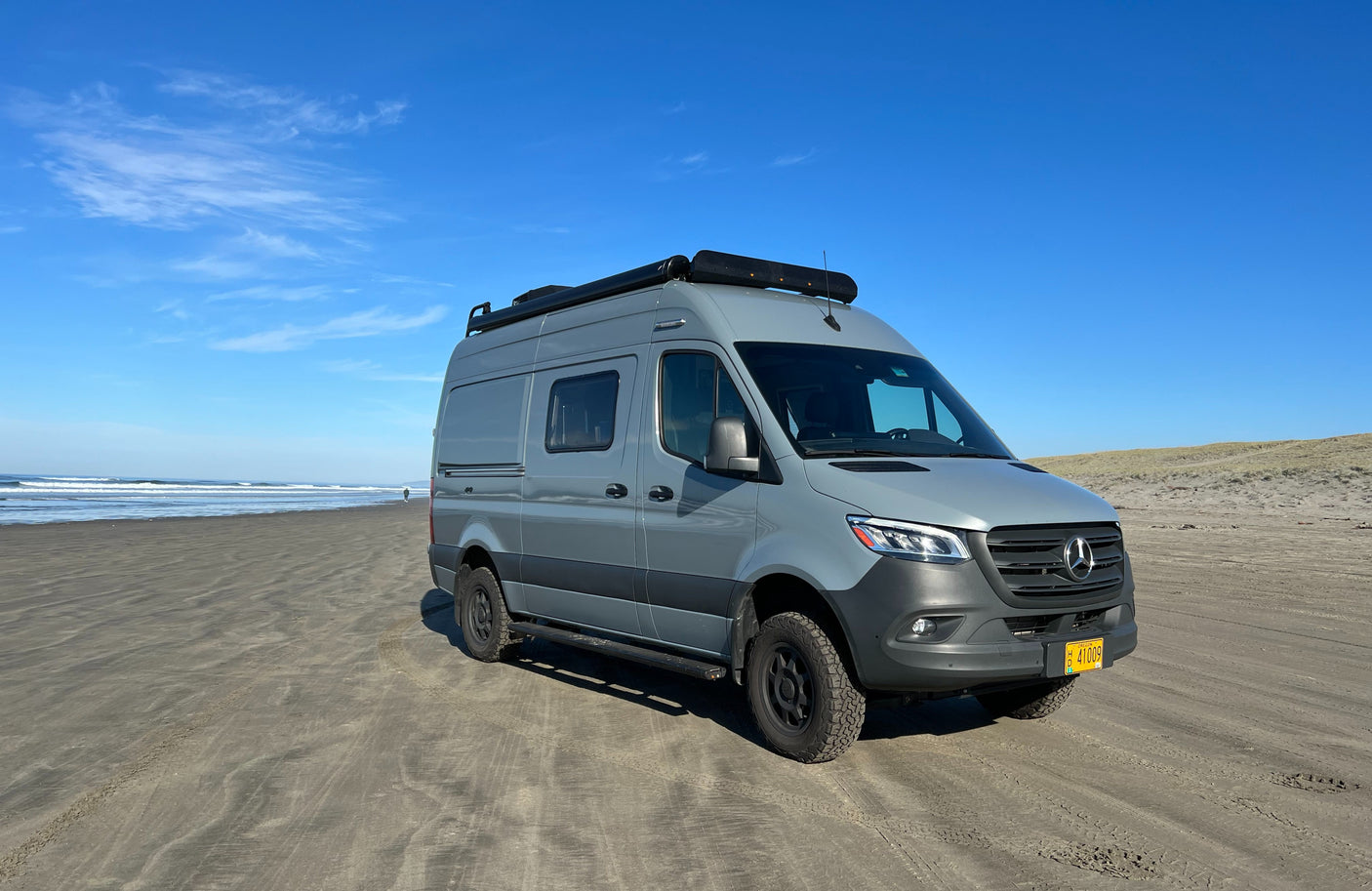 Grey Mercedes Sprinter camper van (RV rental) on a sandy beach by the ocean. A person walks in the distance. Perfect for travel.