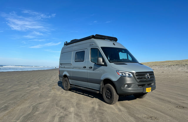 Grey Mercedes Sprinter camper van (RV rental) on a sandy beach by the ocean. A person walks in the distance. Perfect for travel.