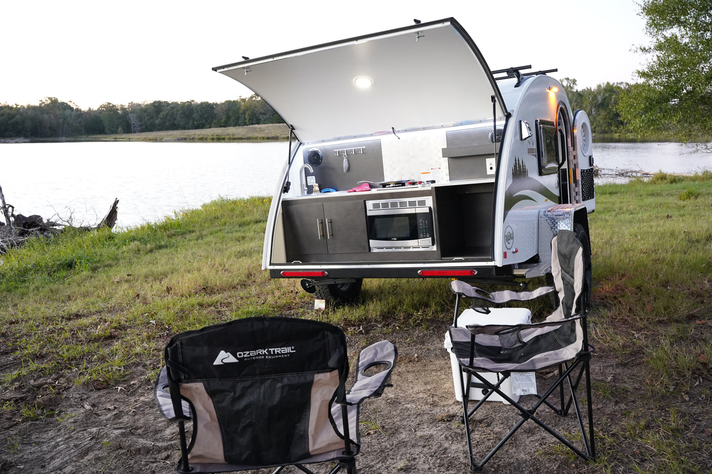 Explore nature with this rv rental! Teardrop camper's outdoor kitchen by a scenic lake, with camping chairs and cooler.