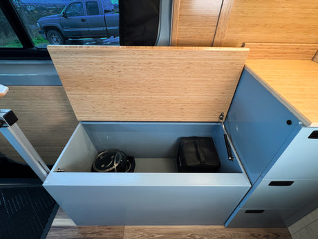 RV rental interior: an open storage compartment revealing kitchenware. Features a bamboo lid, blue cabinetry, and wood-look flooring.