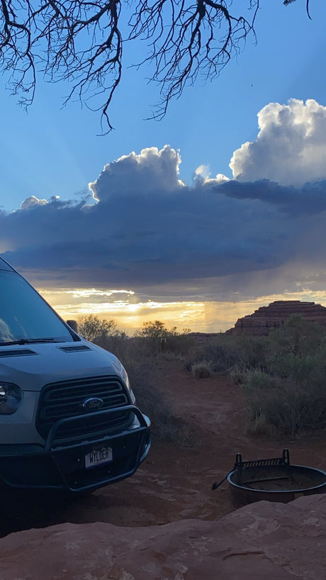 An RV rental Ford Transit camper van parked in a desert at sunset with a fire pit, dramatic clouds, and bare tree branches.