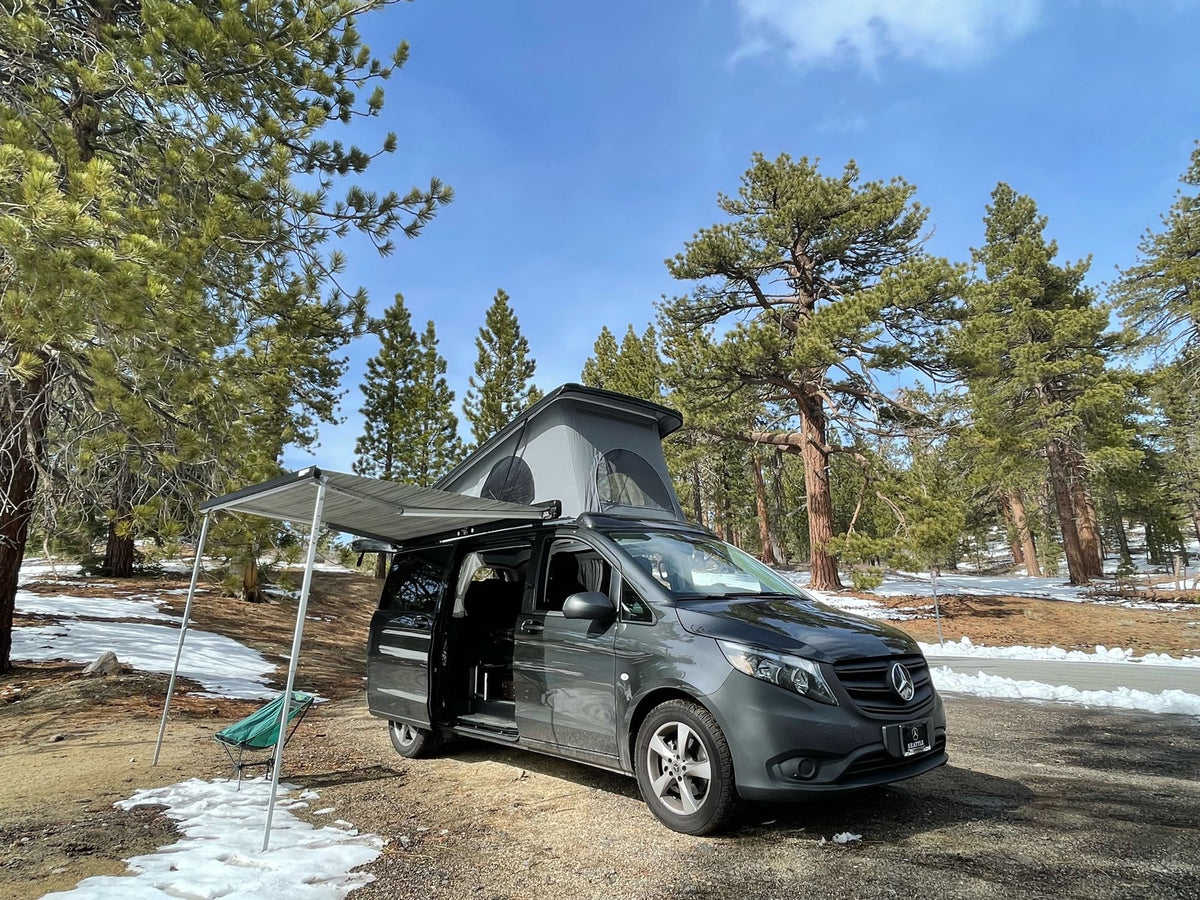 Explore nature in this RV rental: a gray pop-top campervan with an awning set up in a snowy pine forest.|An RV rental: a dark Mercedes-Benz camper van with its pop-top roof open