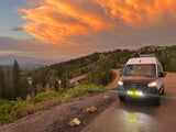 RV rental van on a mountain road at sunset, with glowing orange clouds over forested peaks. Scenic drive, perfect for outdoor adventure.