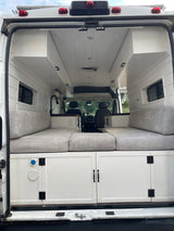 RV rental: Rear view inside a white camper van conversion with a gray cushioned bed, built-in cabinets, and sink.