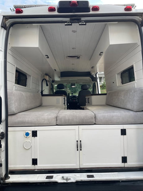 RV rental: Rear view inside a white camper van conversion with a gray cushioned bed, built-in cabinets, and sink.