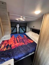 This cozy RV rental features a comfortable bedroom with a vibrant scenic blanket and overhead lighting for your travels.