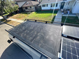 Overhead view of an RV rental's roof with solar panels & perforated rack, parked by houses.
