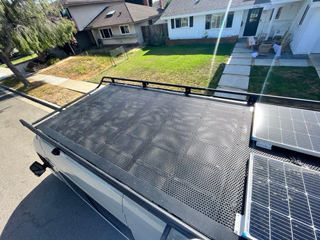 Overhead view of an RV rental's roof with solar panels & perforated rack, parked by houses.