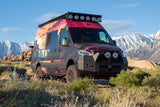 A vibrant custom Sprinter camper van, an ideal rv rental, stands among rocks and brush with snow-capped mountains in the distance.