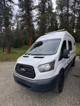 White Ford Transit van, an ideal RV rental, parked on gravel in a lush pine forest.