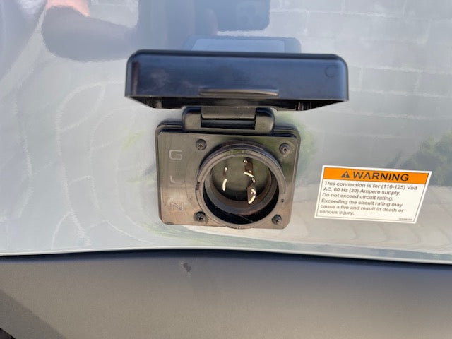 Close-up of an RV rental's 30-amp shore power inlet with its cover open, revealing three prongs. A warning label details 110-125V AC supply.