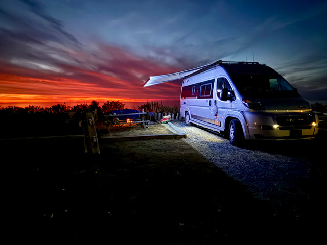 A modern RV rental with an extended awning illuminates a beach campsite at sunset, showcasing vibrant red and orange skies.