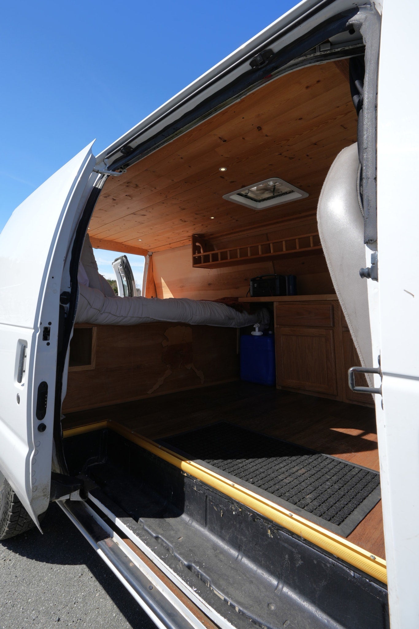 View inside an RV rental camper van: comfortable bed, wood-paneled walls/ceiling, kitchen area, and open side door. — photo 7