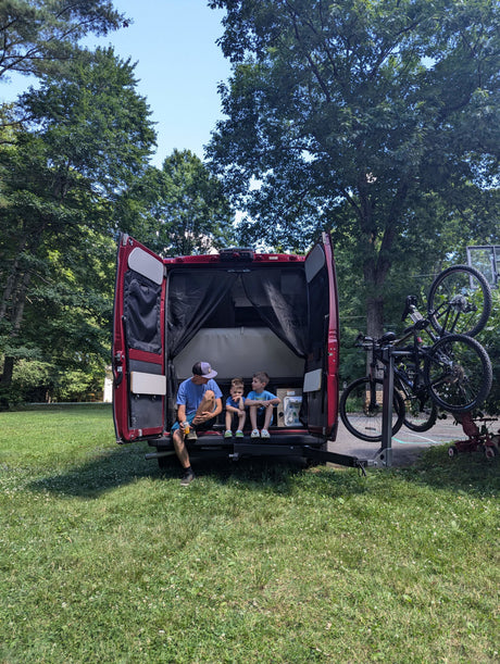 Enjoying the rv rental van! A man and two boys relax in the open back, surrounded by nature with bikes mounted for adventure.