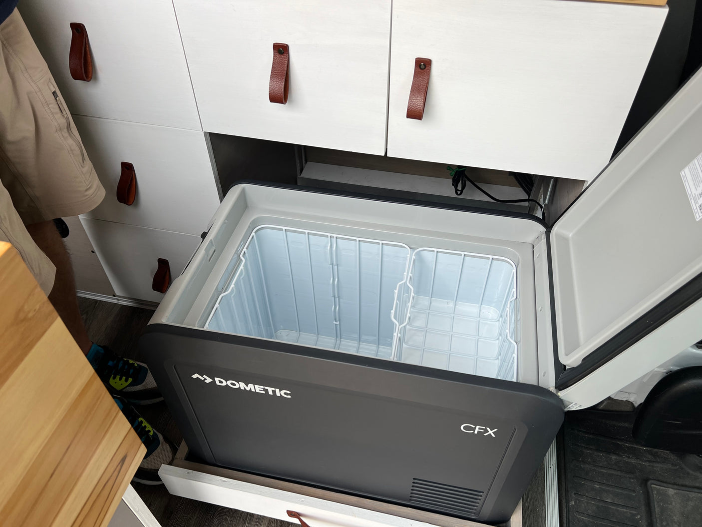 Open Dometic CFX portable fridge in an rv rental camper van, showing wire baskets & white cabinets, ready for travel.