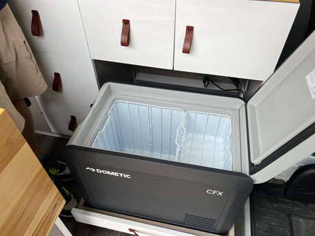 Open Dometic CFX portable fridge in an rv rental camper van, showing wire baskets & white cabinets, ready for travel.