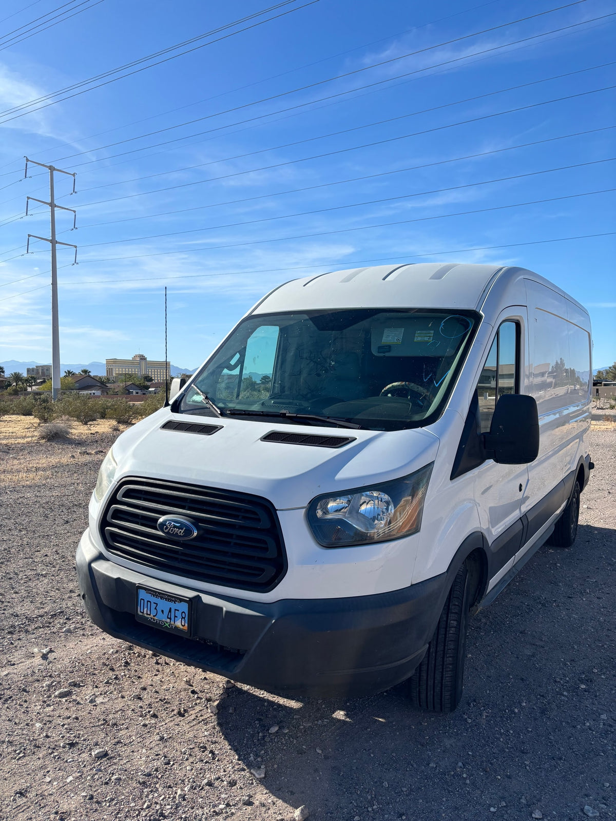 White Ford Transit cargo van at a desert lot. Power lines and distant buildings are in the background. Potential for an rv rental conversion. — photo 4